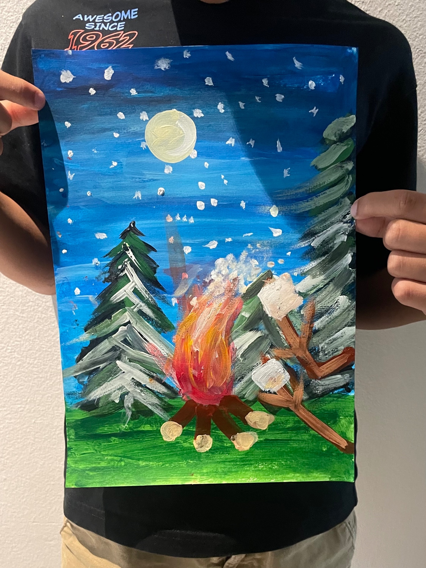 Student holding an acrylic painting of a snowy campfire scene under a full moon